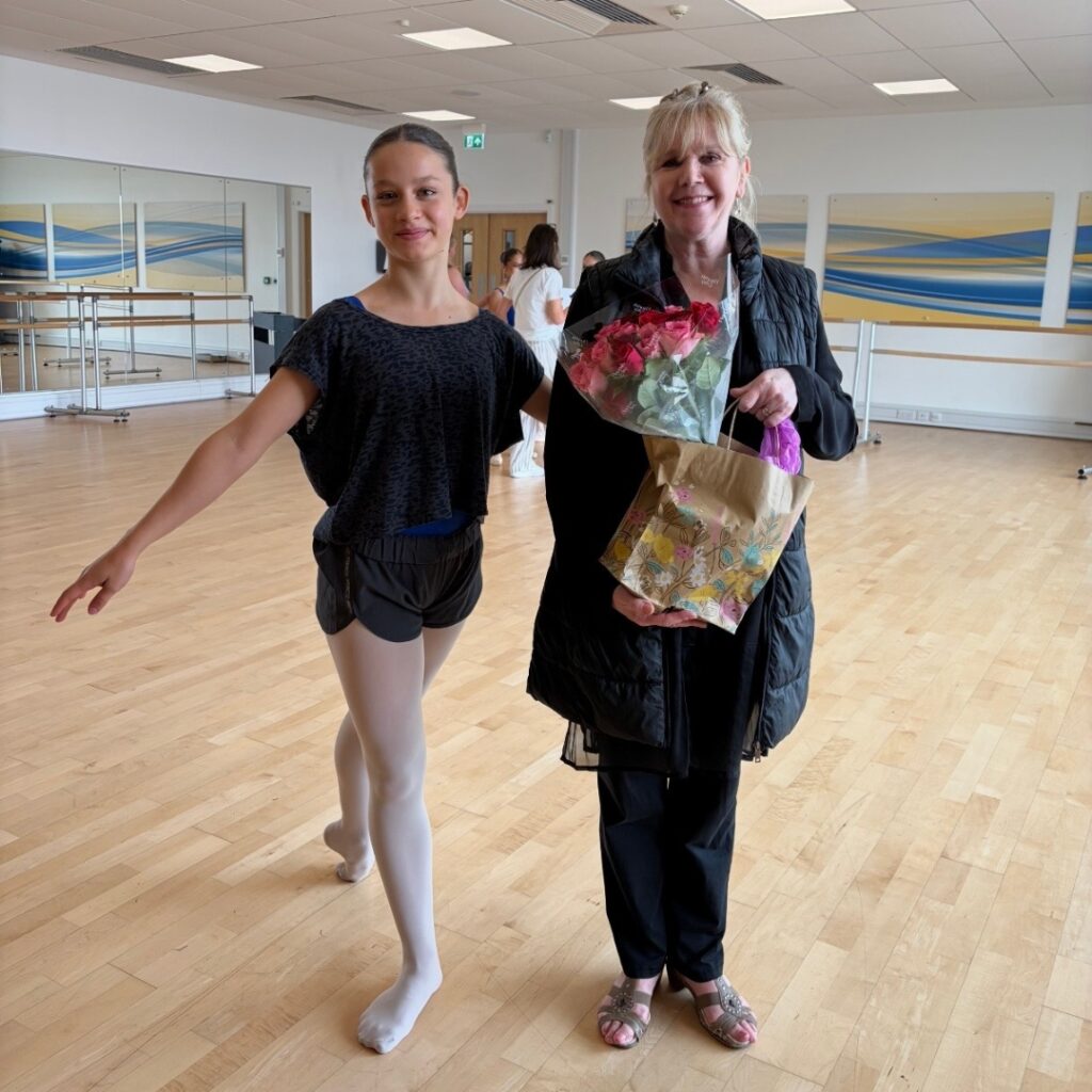 Mid associate students share what they love about the associate programme Katie wears a black t-shirt and shorts over pink tights with her left leg in tendu back. She stands next to karen paisey, who wears a black coat and holds a bouquet of flowers.