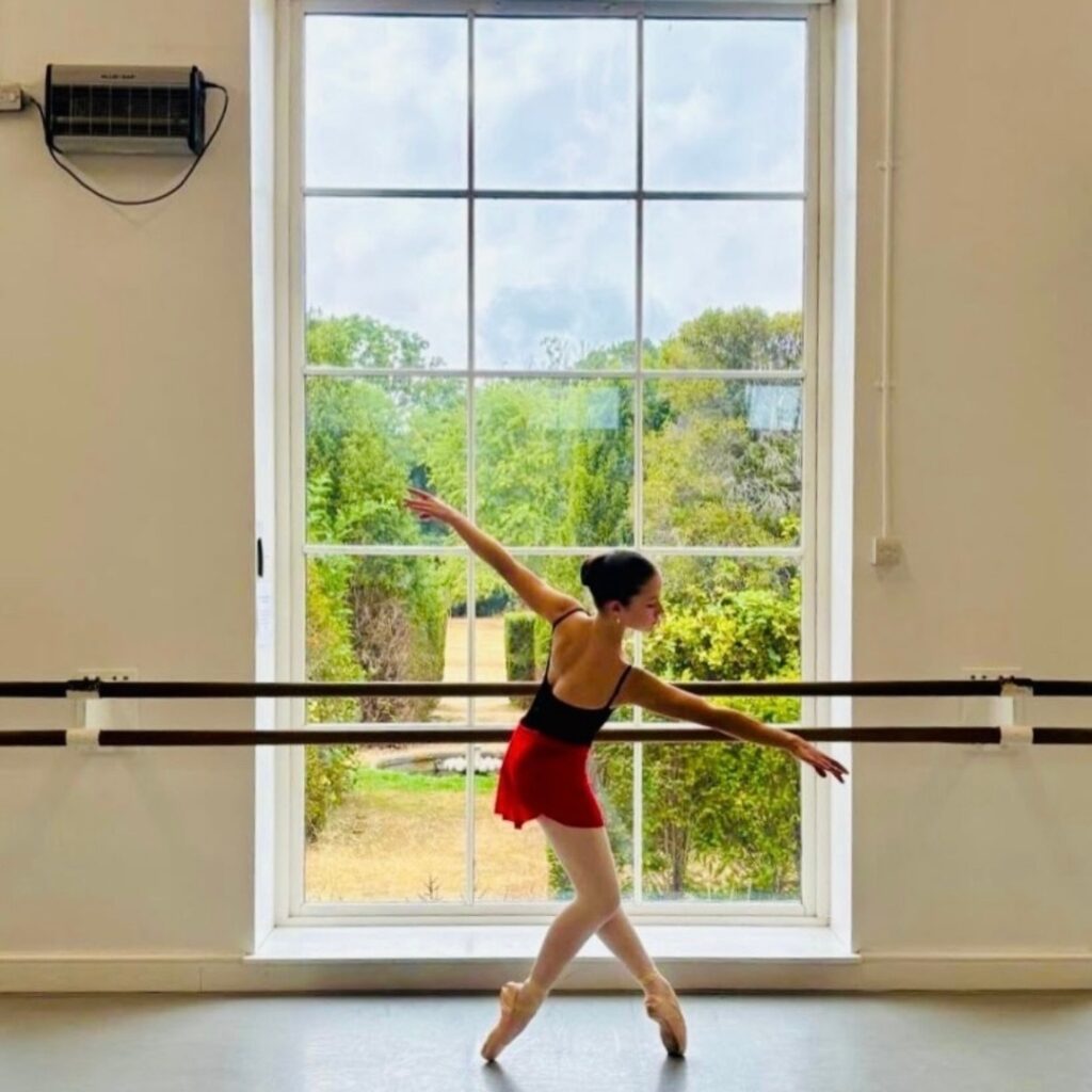 Mid associate students share what they love about the associate programme Katie balances on pointe in a white lodge studio in front of a window overlooking a green garden. Her left foot is extended front and her right leg is bent to the back. Her right arm is on a diagonal down to the front with her left in a line up to the back. Her back faces the camera with her face in profile.
