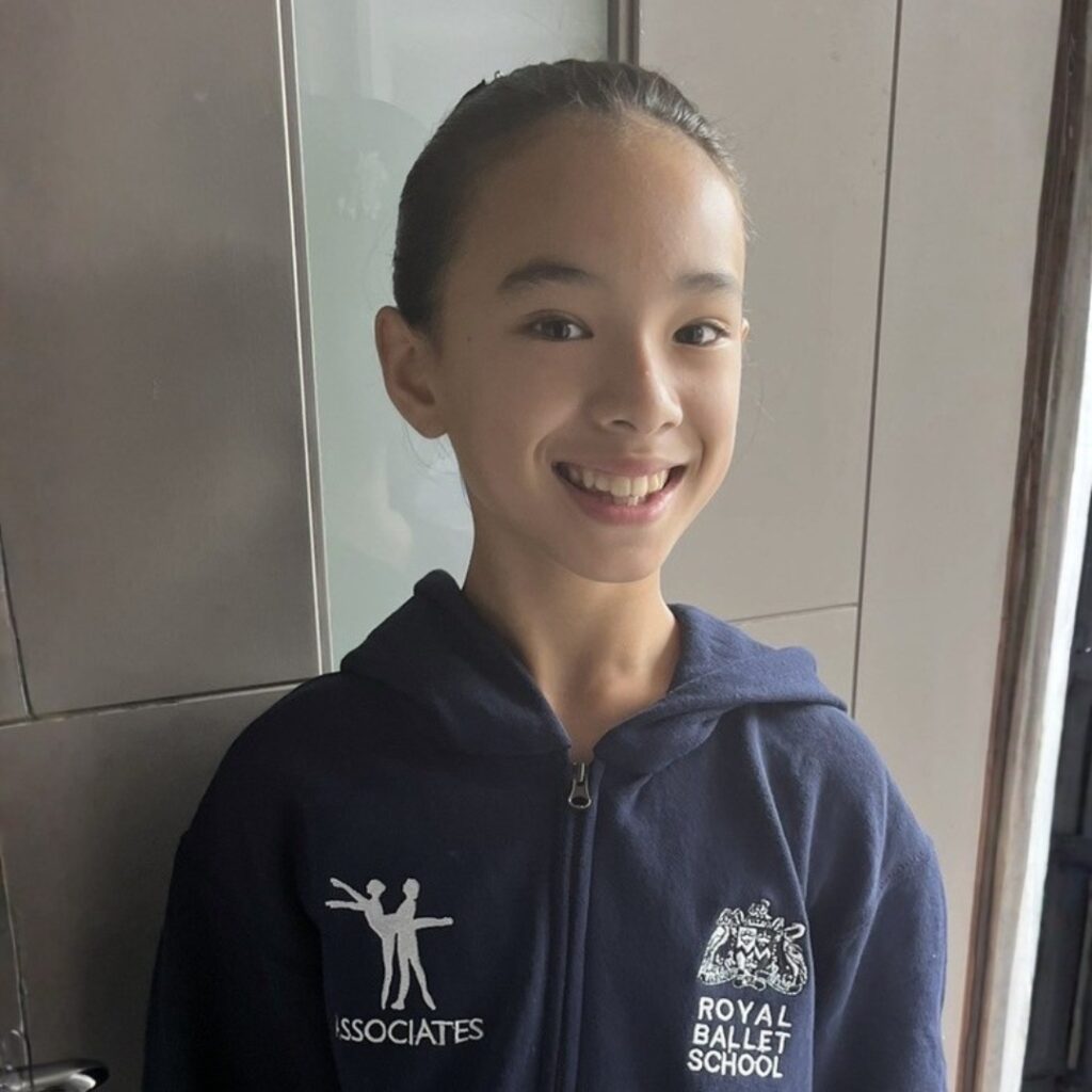Mid associate students share what they love about the associate programme Maisie smiles at the camera in front of a white wall. She wears an associates tracksuit with her dark hair in a bun.