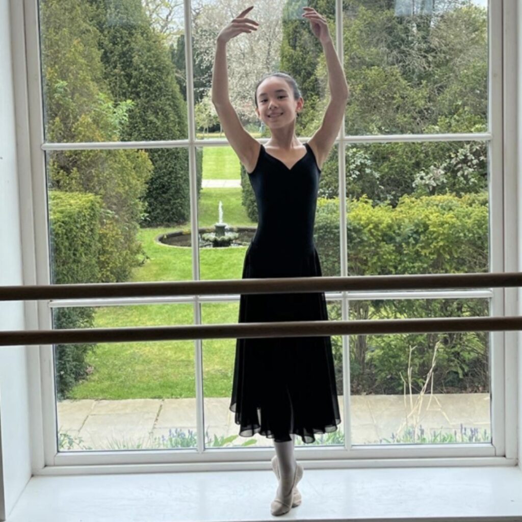 Mid associate students share what they love about the associate programme Maisie poses in fifth position on releve in flat shoes in front of a window at white lodge that overlooks the green gardens and fountain. She wears a black leotard and a long black skirt.