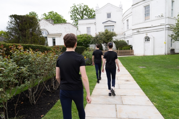 A day in the life of a full-time student at the royal ballet school Three students in black shirts and trousers walk outside of the royal ballet school - white lodge on a stone path cutting through grass.