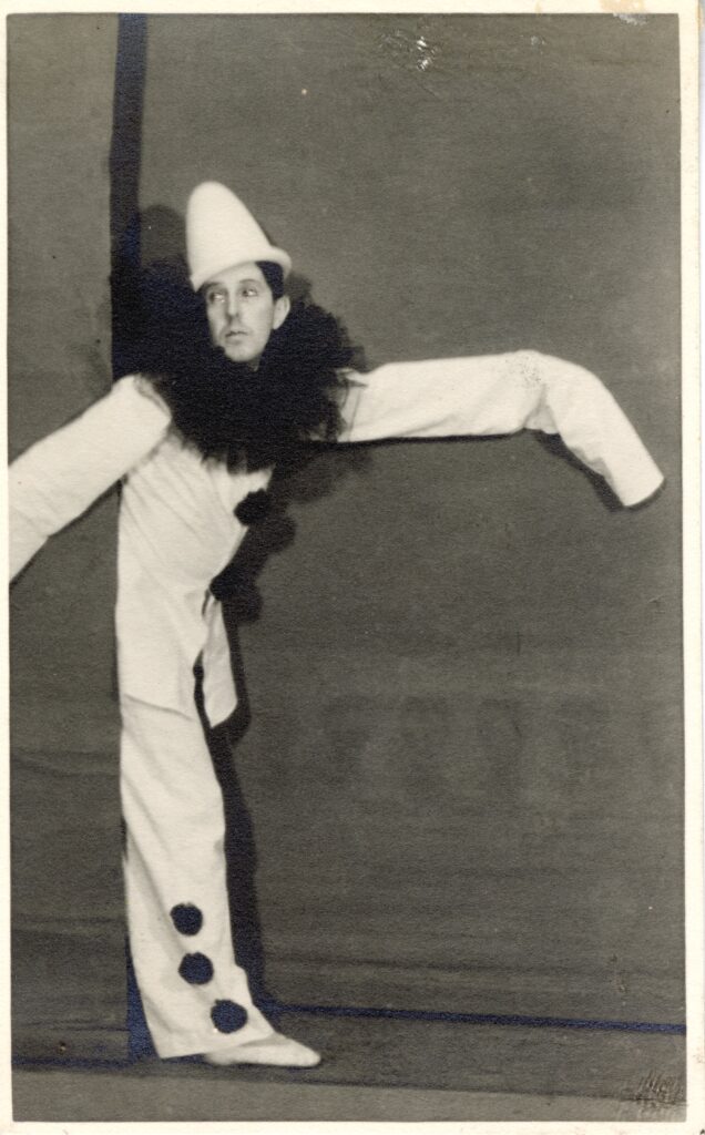 100 stories: frederick ashton Ashton wears a black and white costume with a feather ruff, walking on stage from behind a curtain.