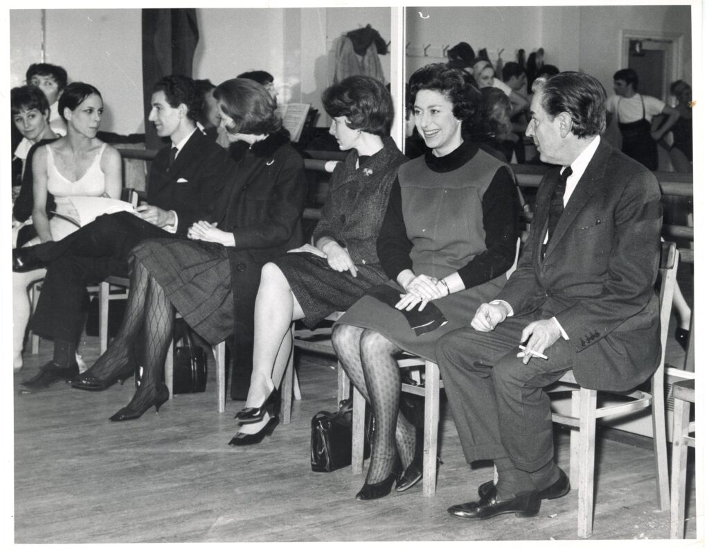 100 stories: frederick ashton Ashton sits next to princess margaret watching a ballet class. They sit in front of a mirror and smile as they watch the class.