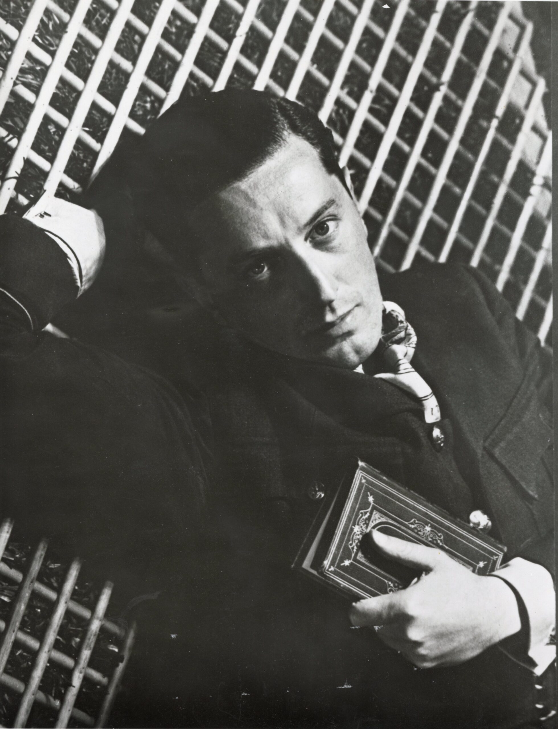 100 stories: frederick ashton Ashton lays down with his right hand behind his head and his left hand holding a book on his chest. He wears a black suit and looks towards the camera intently. Part of the royal ballet school's 100 stories series.