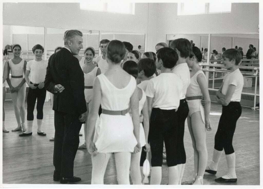 100 stories: frederick ashton Ashton teaches a class at white lodge in 1970. He stands in front of a group of boys and girls and wears a black suit. The girls are in white tunics with belts, and the boys wear white shirts, black capri length tights and black shoes.