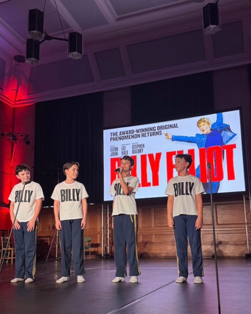 Year 8 student rafferty on starring as the titular role in billy elliot the musical The four billys stand on stage with the musical's poster on a screen behind them. They wear navy trousers with yellow stripes running lengthwise down the sides, white ballet shoes and white t-shirts with black text that reads 'billy'