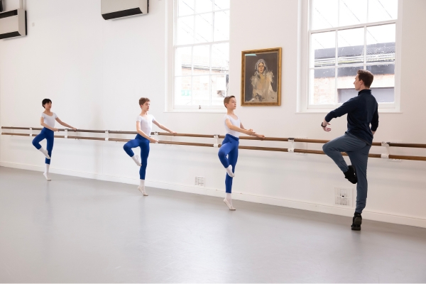 A day in the life of a full-time student at the royal ballet school Three boys stand with their left hands on the barre with their right legs in passe. Their left leg is on releve, and their right arms are in first position. In front of them, a male teacher mirrors the movement.