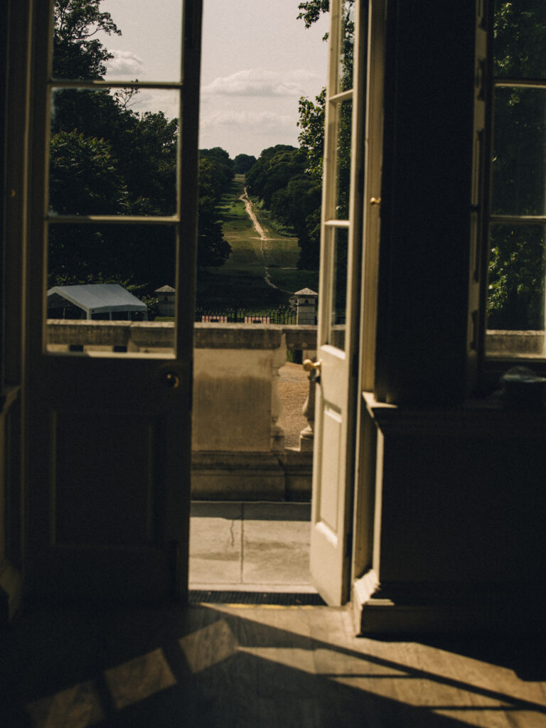 A photo from inside white lodge looking outside to the queen's ride cutting a straight line through the tree-lined grass. In the foreground, a door frame with glass panels frames the photo.