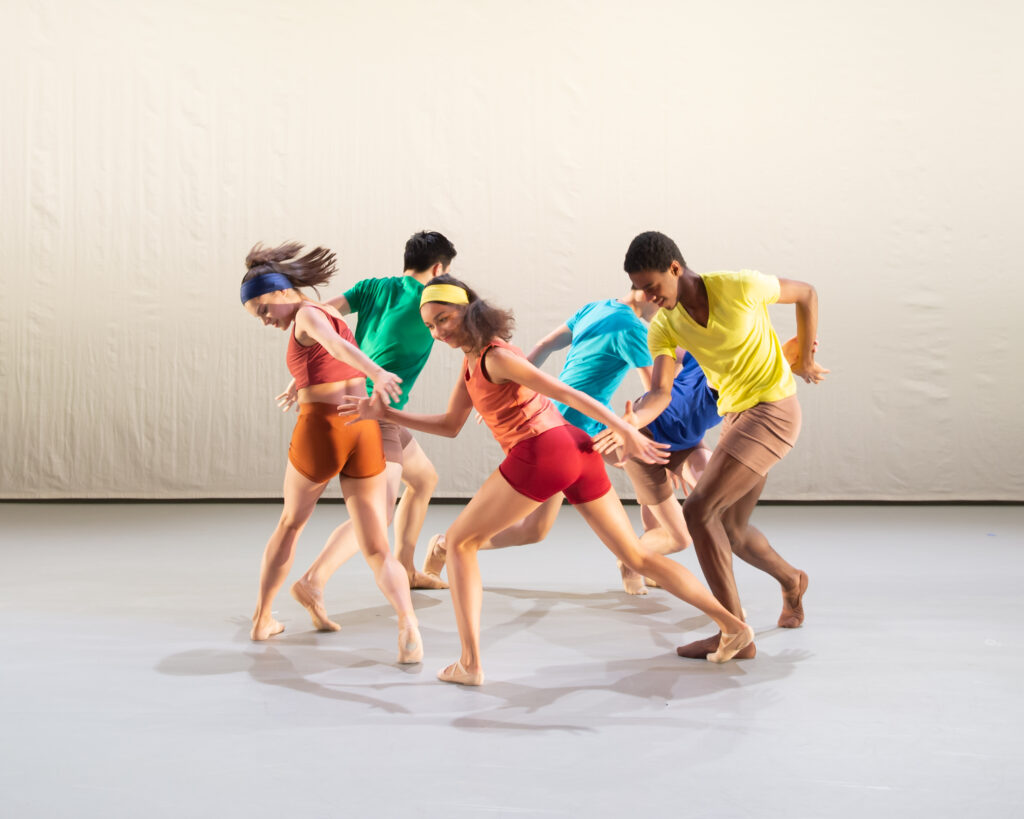 Denilson almeida's 2019 choreography the middle of nowhere, performed at white lodge. A group of six young dancers wear a rainbow of different coloured shirts, shorts and headbands. They hold hands and run in a circle. They are barefoot, and long hair is pulled into a ponytail.