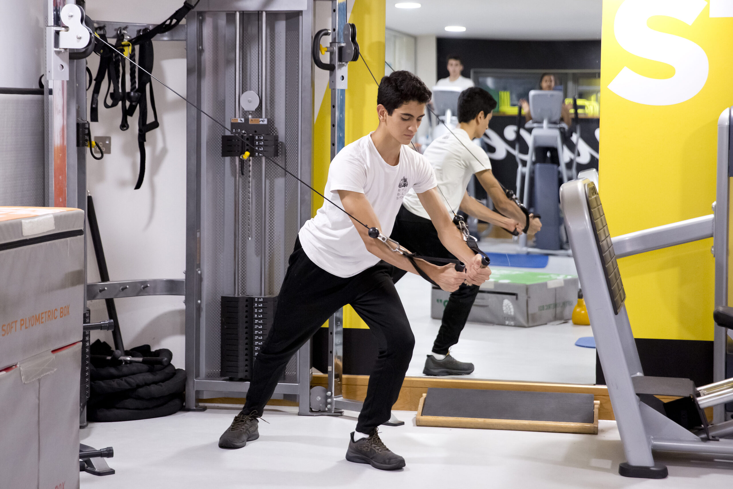 Denilson wears a white royal ballet school t-shirt and black trousers with brown trainers. He pulls a resistance machine down and forward.