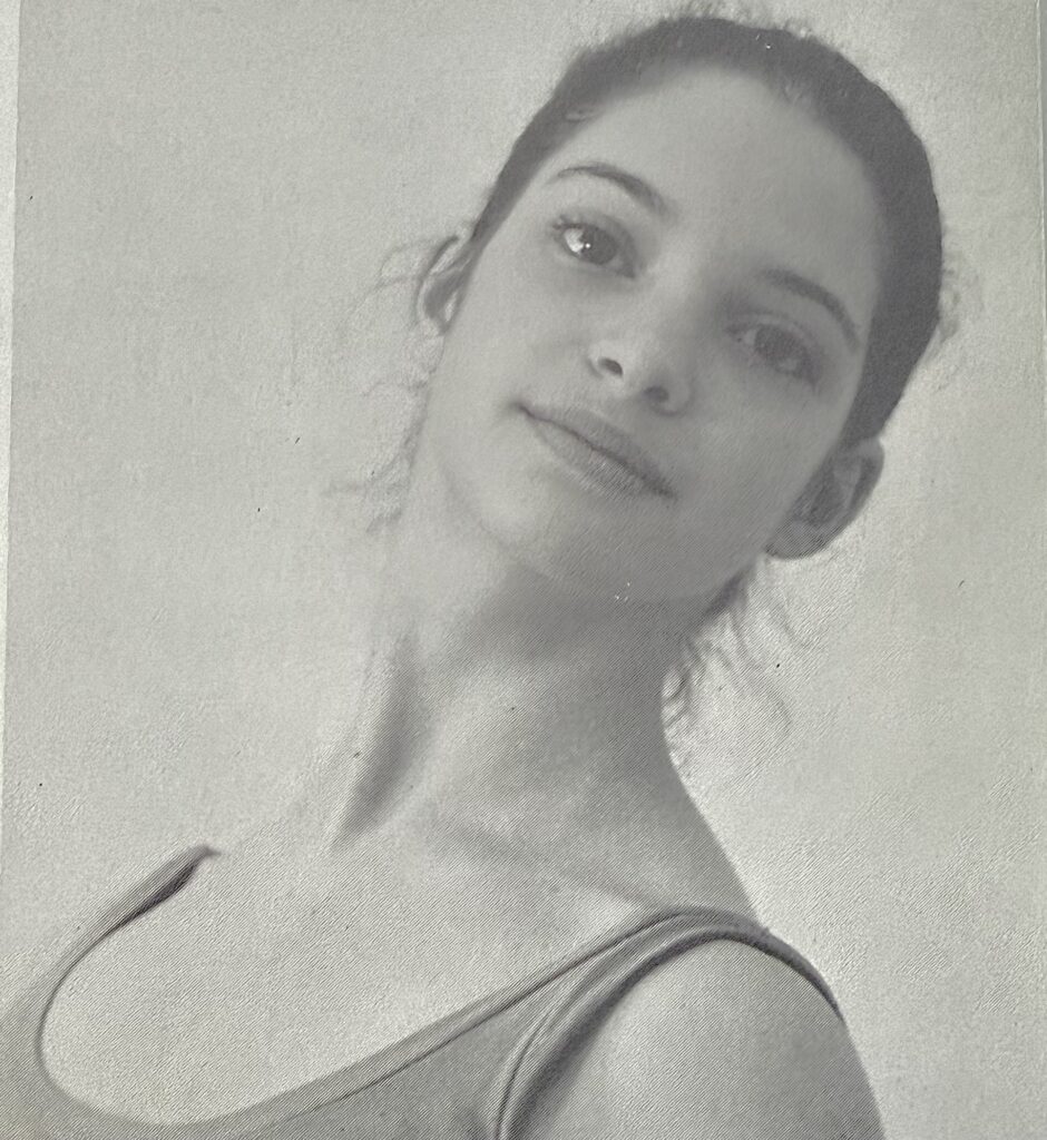 A black and white school portrait of rachel at white lodge for our 100 stories series. Her hair is in a high bun with wisps of hair framing her face. She smiles softly at the camera.