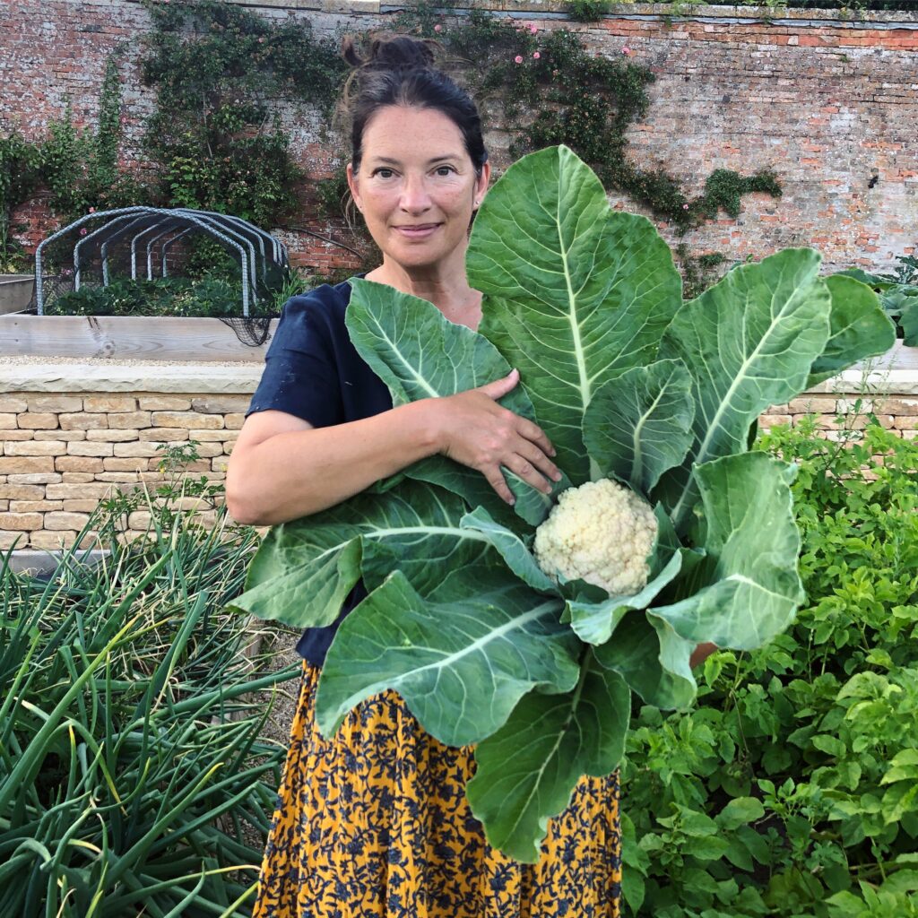 Rachel holds a cauliflower with large green leaves at broadwell. Her hair is tied back, and she smiles softly to the camera.