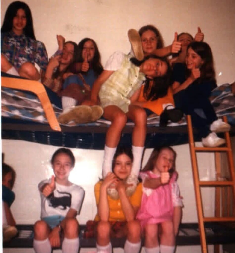 A group of 11 girls pile onto two bunk beds, smiling and posing goofily at the camera. Rachel sits on the middle of the bottom bunk.