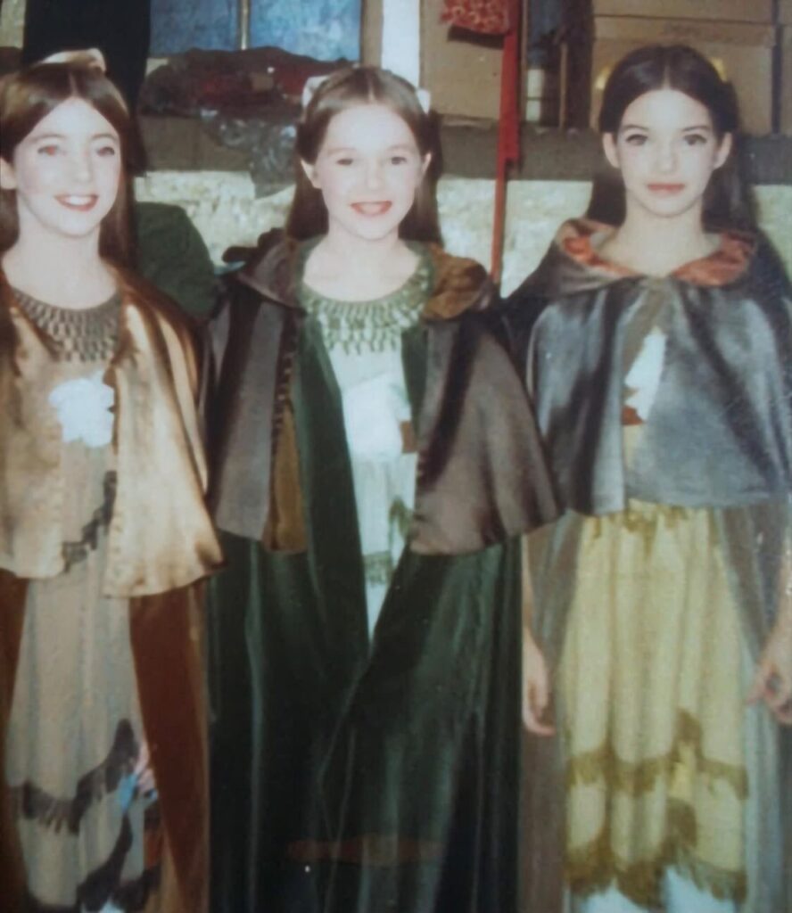 Three girls dressed in early 20th century costumes smile at the camera, backstage at the nutcracker.